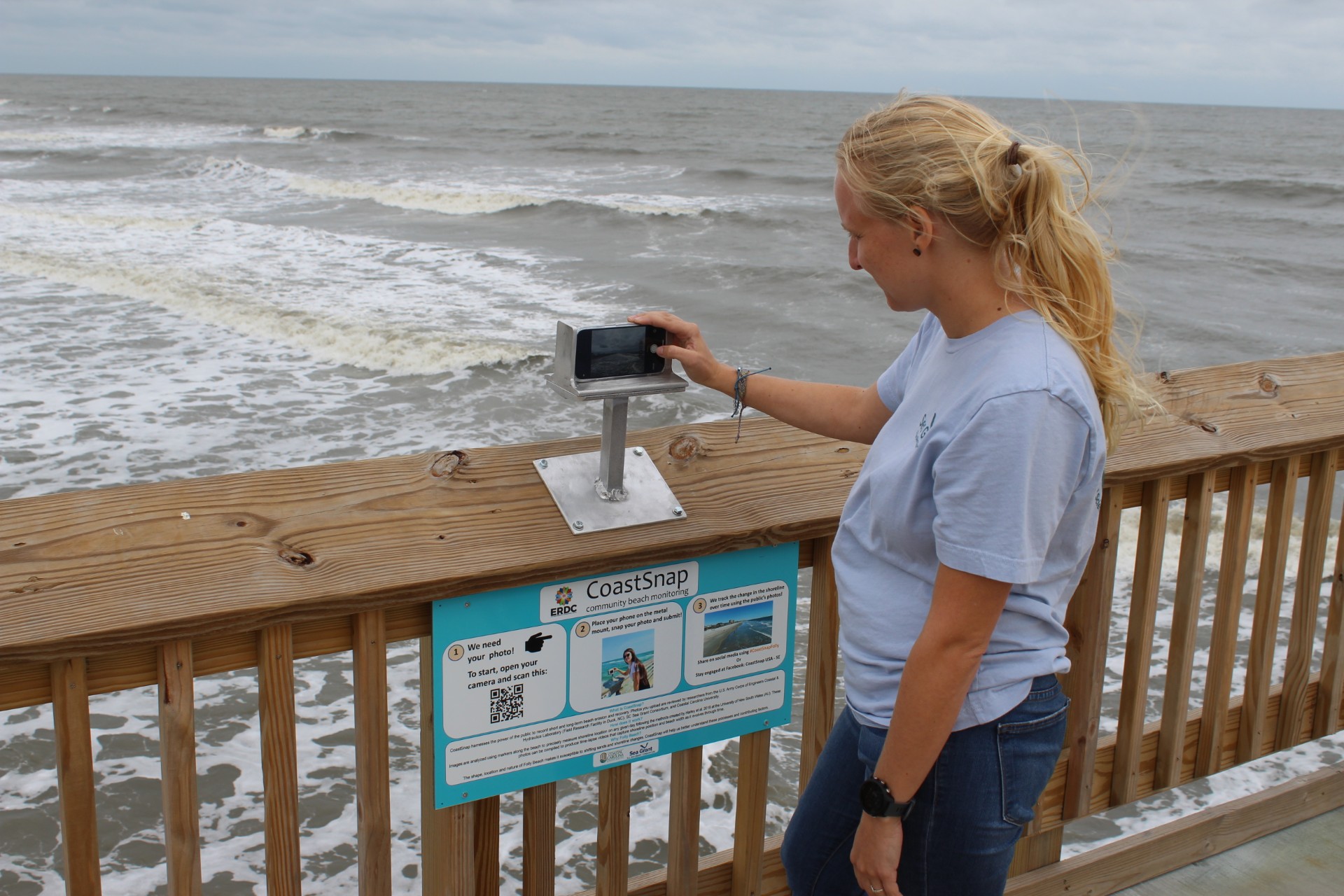 person standing on a pier over the ocean holding a phone in a metal box overlooking the water
