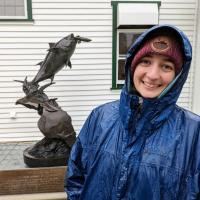 A young woman in a blue rain coat and maroon beaned stands in front of a statue of a tuna at the in front of a statue at the Catalina Tuna Club.