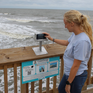 person standing on a pier over the ocean holding a phone in a metal box overlooking the water