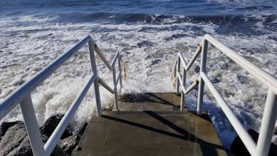 Rough waves crash into cement stairs lined with white railings that lead into the ocean. 
