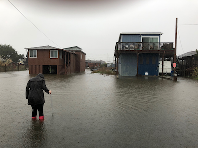 Person walking in a flooded street towards a couple of flooded houses.
