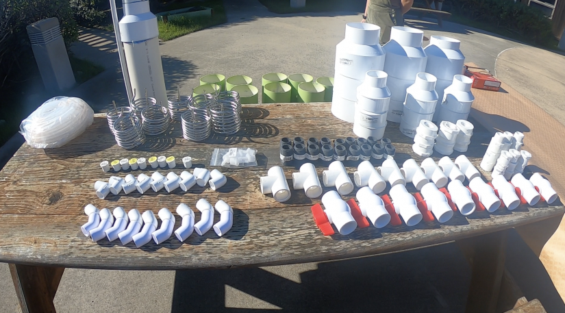 A picture of various parts for building multiple CLASH units. PVC parts and fittings, heat coils, valves, and air tubing is organized grouped by part on an outdoor table at Moss Landing Marine Lab.
