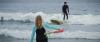 Surfer girl walks into the water carrying a surfboard, while a young man surfs a paddle board on a wave.